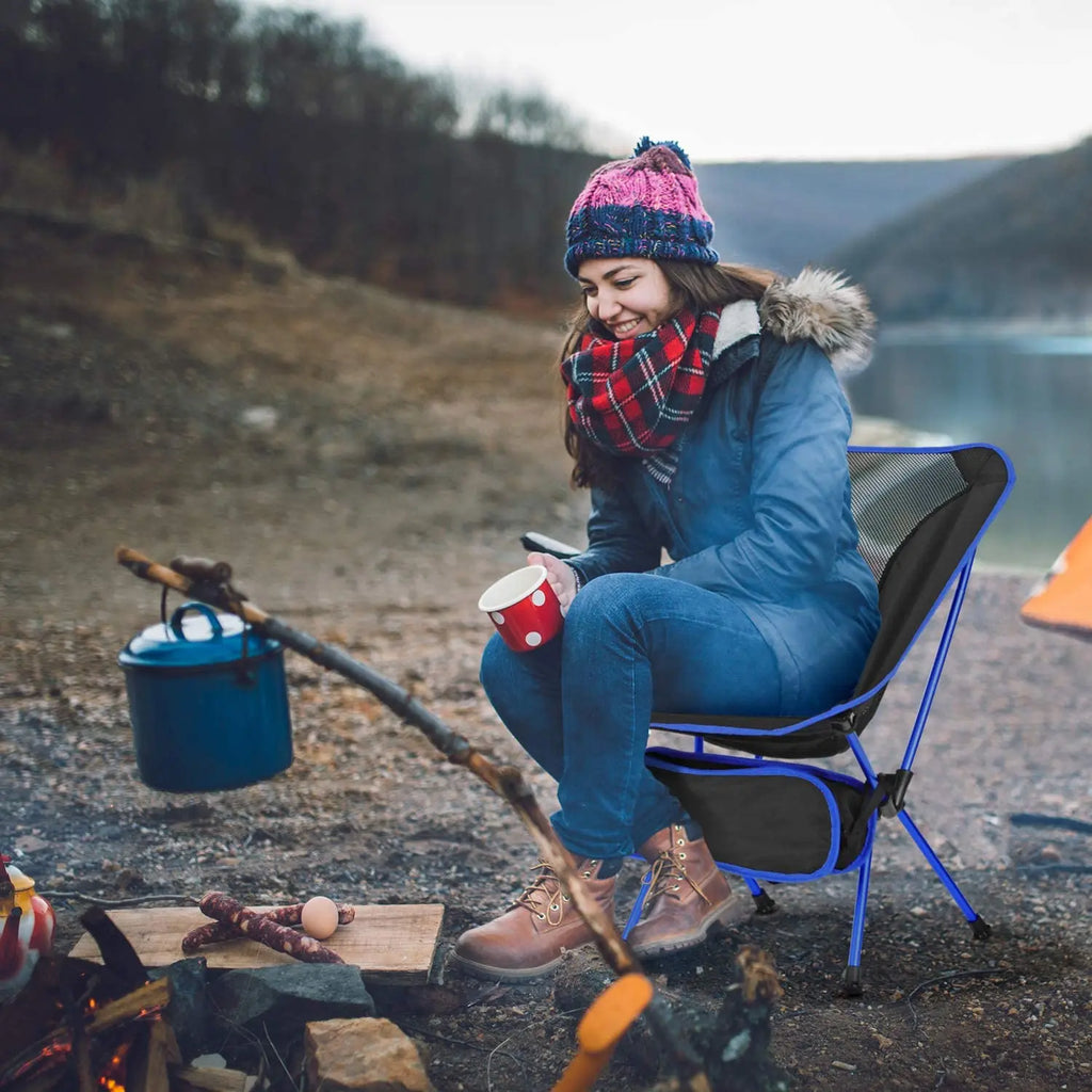 A woman sitting on a portable blue camping chair by a campfire near a lake.