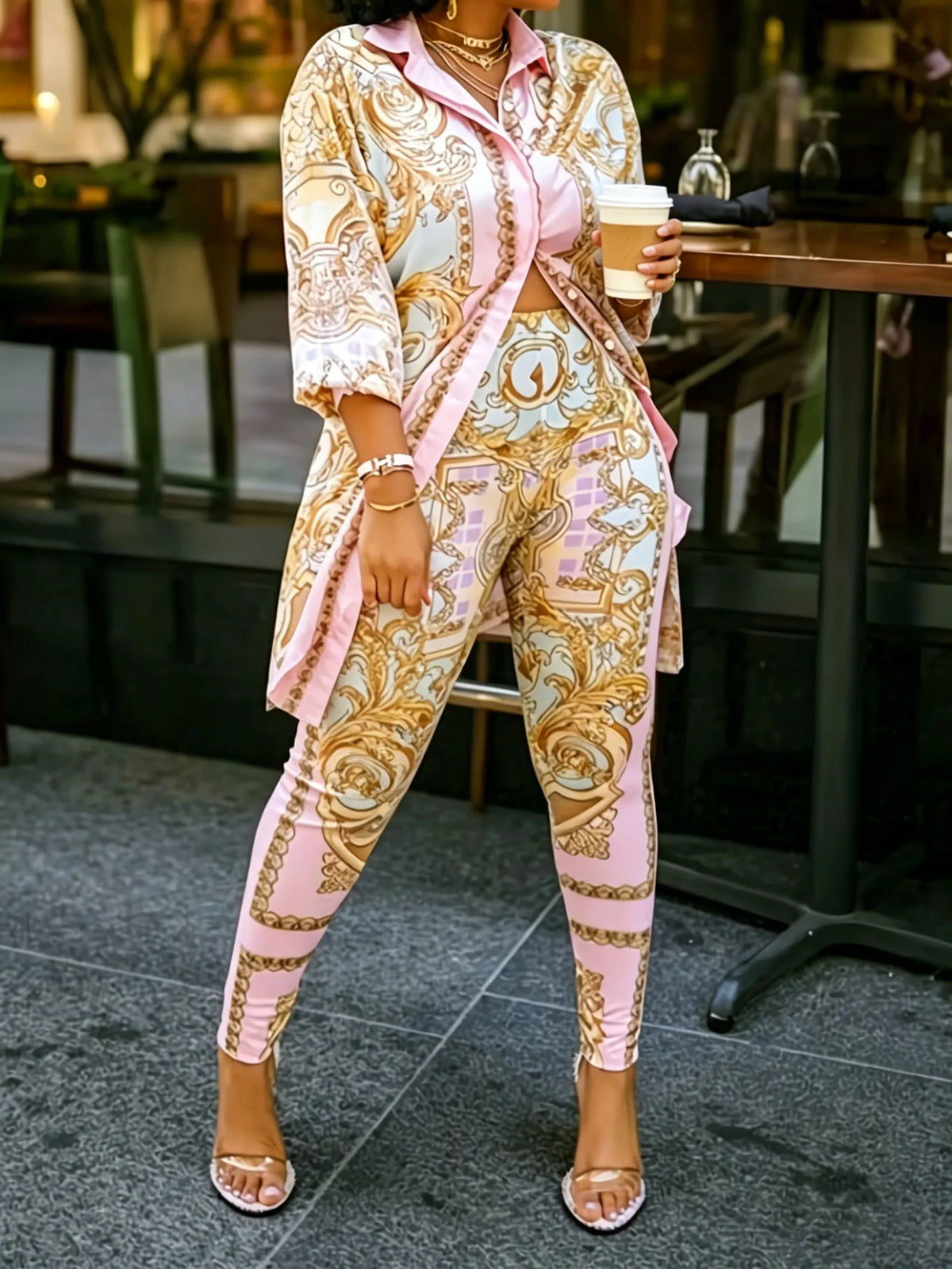 A woman wearing a pink and gold ornate print two-piece shirt and pants set, holding a coffee cup.