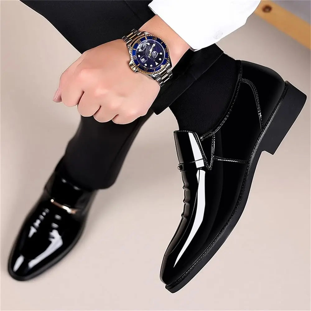 A man wearing a wristwatch and black formal trousers, presenting a pair of black glossy patent leather loafers.