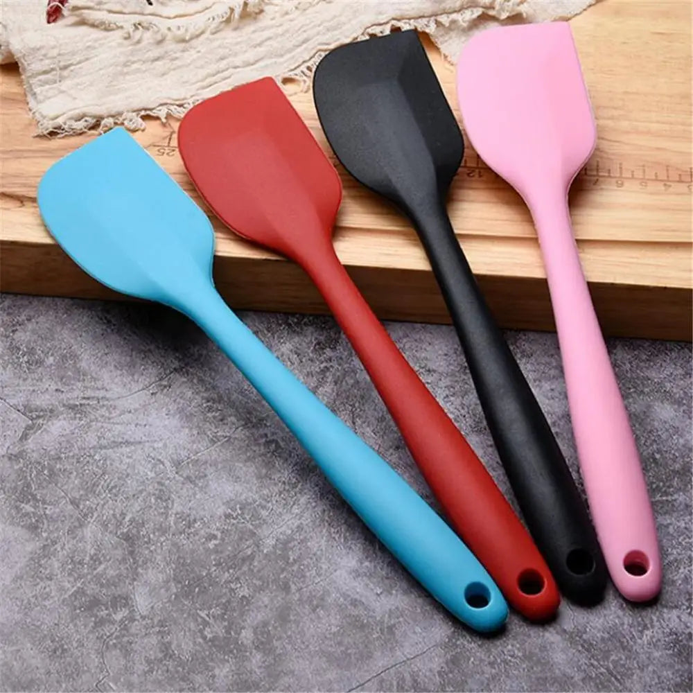 Four silicone spatulas in blue, red, black, and pink colors resting on a wooden board.