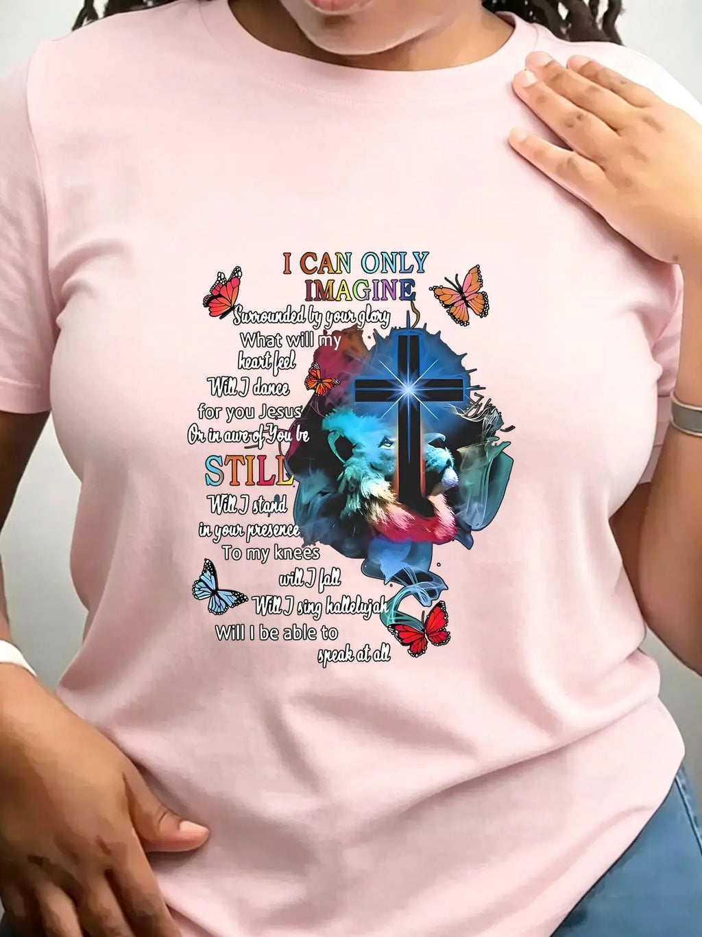 A light pink short-sleeved crew neck t-shirt with a religious graphic print featuring a cross, lion, butterflies, and lyrics