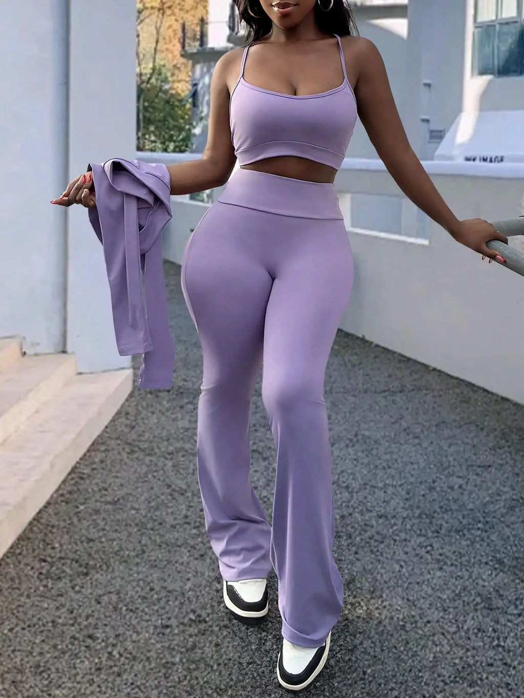 A woman in a purple sports bra and matching flared trousers, holding a long-sleeve zip-up top.