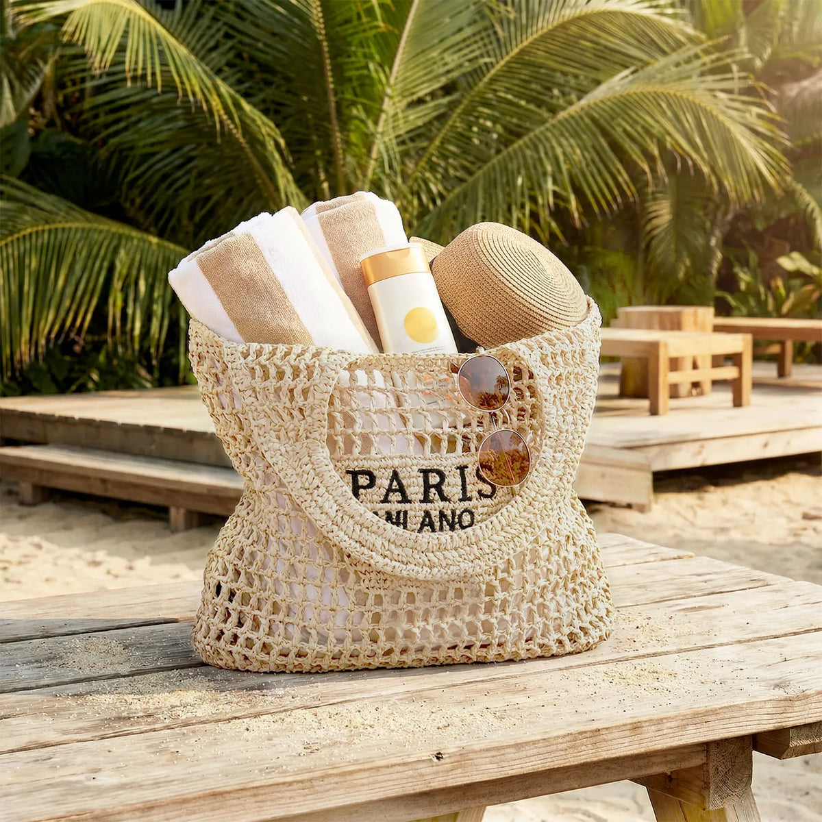 A beige woven mesh beach bag filled with towels and sunscreen on a wooden table at the beach.