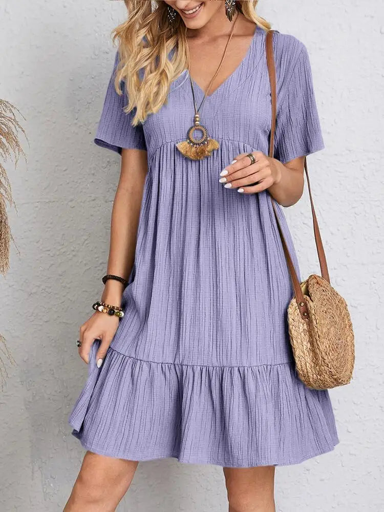 A woman wearing a short-sleeved light purple V-neck textured mini dress with a tiered ruffle hem and a round straw crossbody