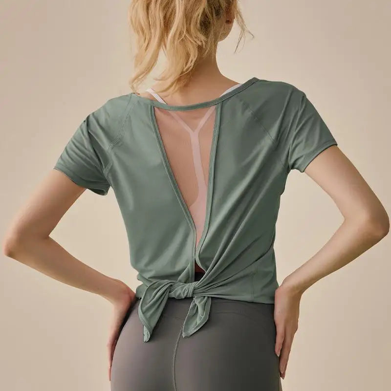 Back view of a woman in a sage green short-sleeve yoga shirt with a mesh back and tie hem detail