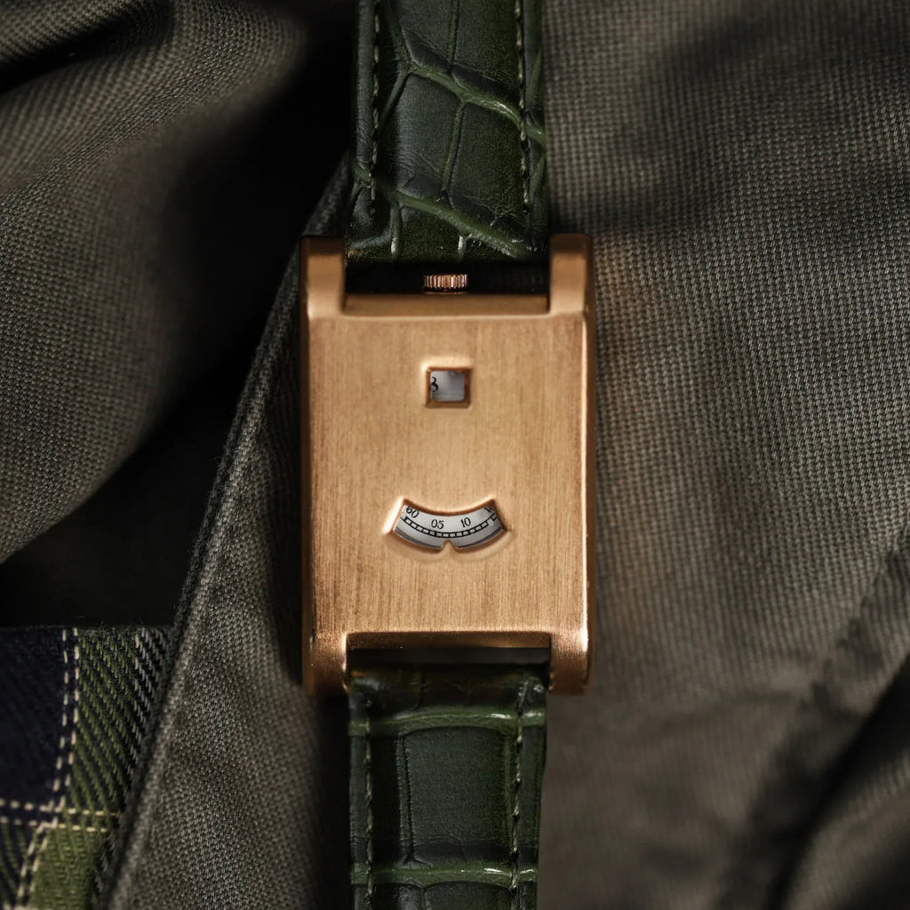A close up of a rose gold watch with a green alligator print leather strap against a fabric backdrop.