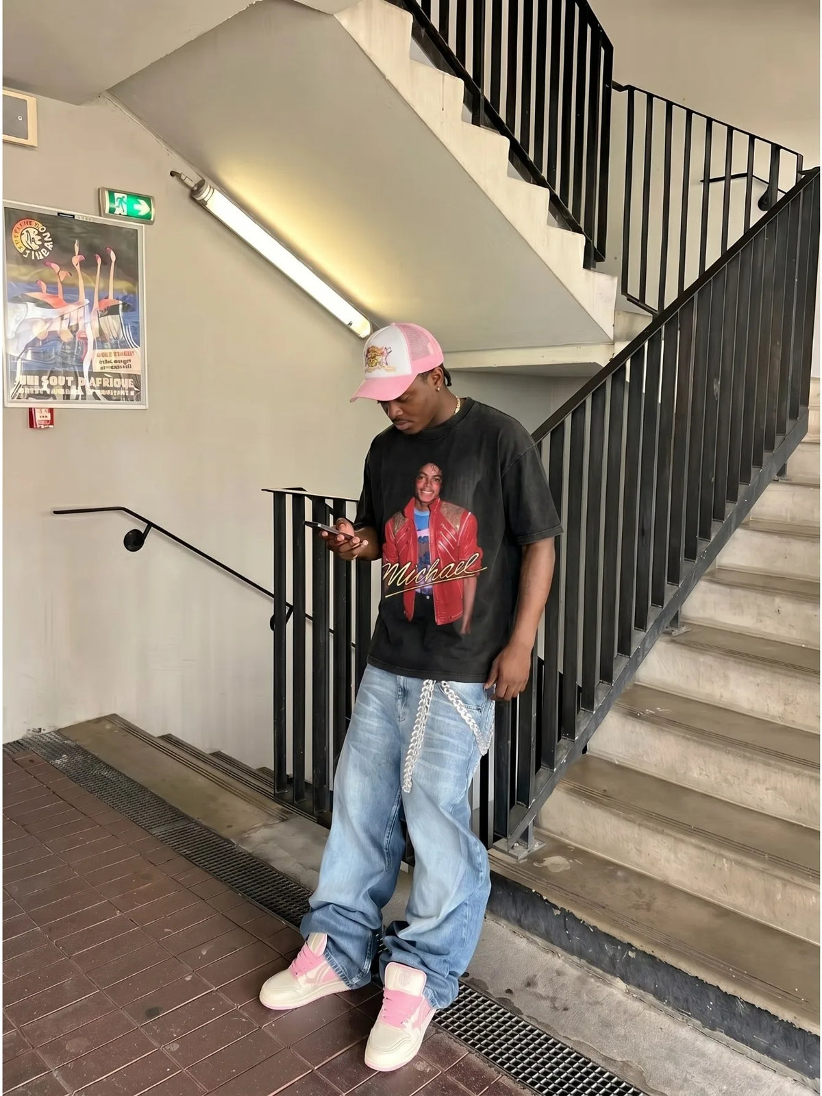 Man standing on stairs holding a phone, wearing a black graphic t-shirt, baggy jeans, sneakers, and a pink baseball cap