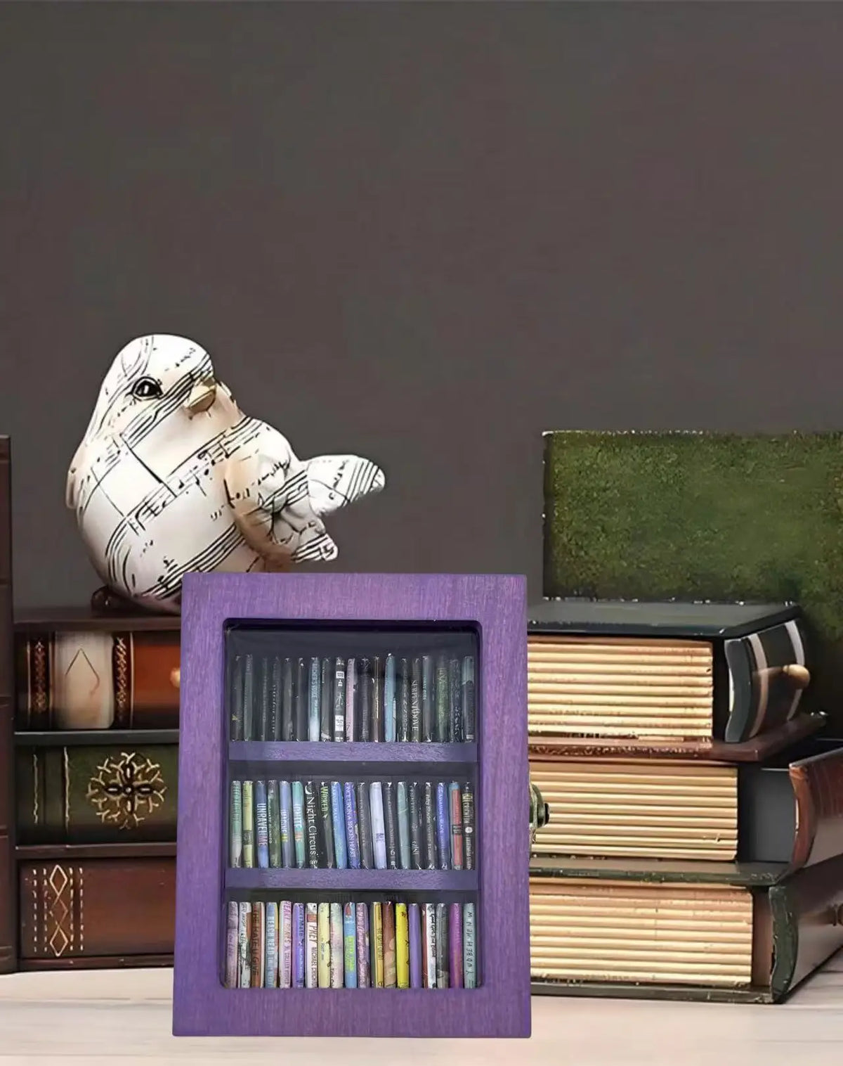 A small purple wooden bookshelf filled with miniature books, displayed on a desk next to a decorative music-themed bird
