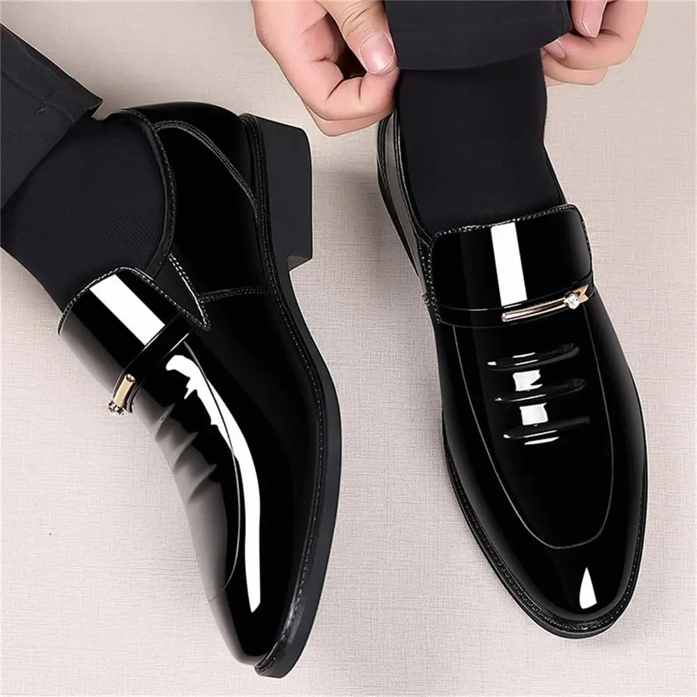 Pair of glossy black patent leather mens formal loafers with gold metal detail worn by a man.