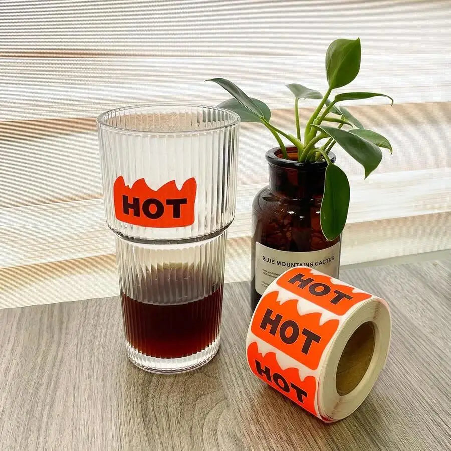 A glass with a flame-shaped HOT sticker and a roll of stickers next to a potted plant
