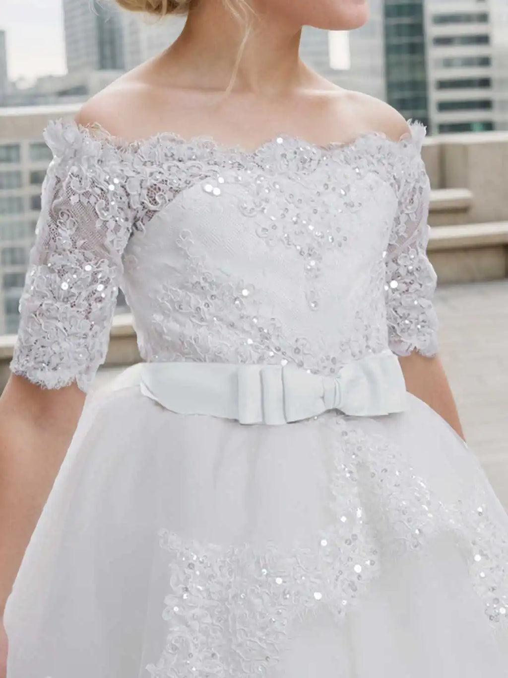 Close up of the bodice of a white off-shoulder lace and sequin flower girl dress with a bow at the waist.
