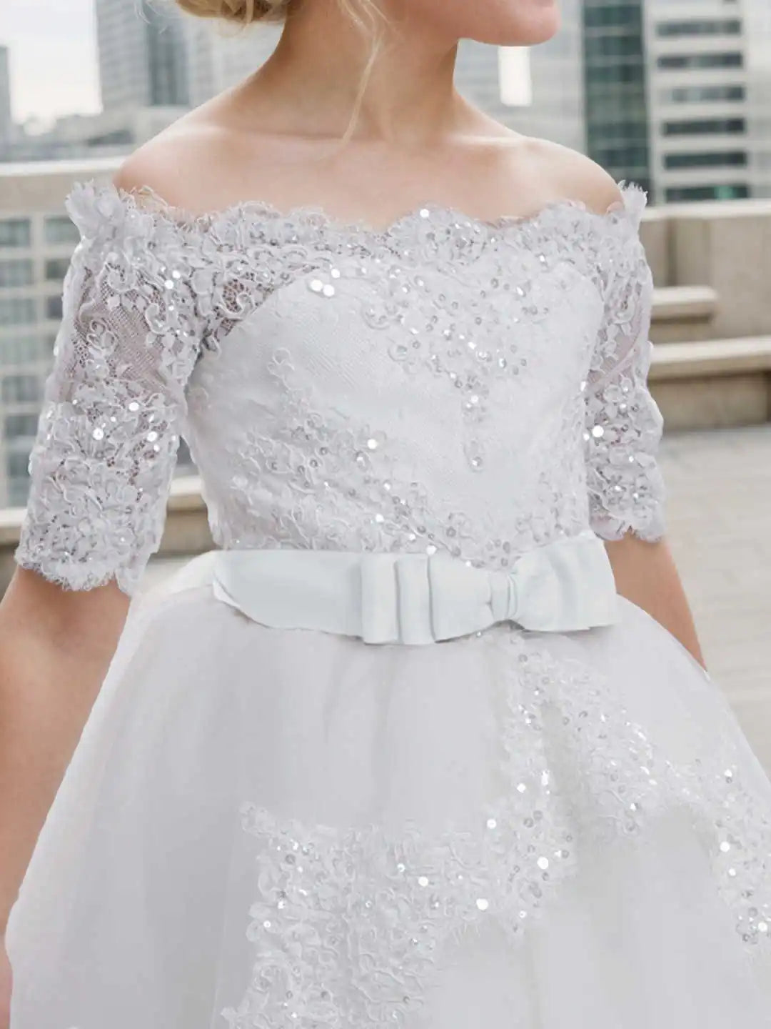 Close up of the bodice of a white off-shoulder lace and sequin flower girl dress with a bow at the waist.