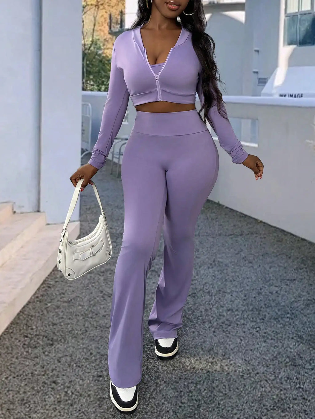 A woman wearing a purple long-sleeve zip-up top and matching flared trousers, carrying a white shoulder bag.