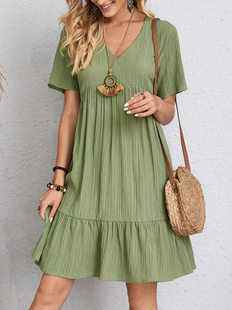 A woman wearing a short-sleeved green V-neck textured mini dress with a tiered ruffle hem and a round straw crossbody bag.