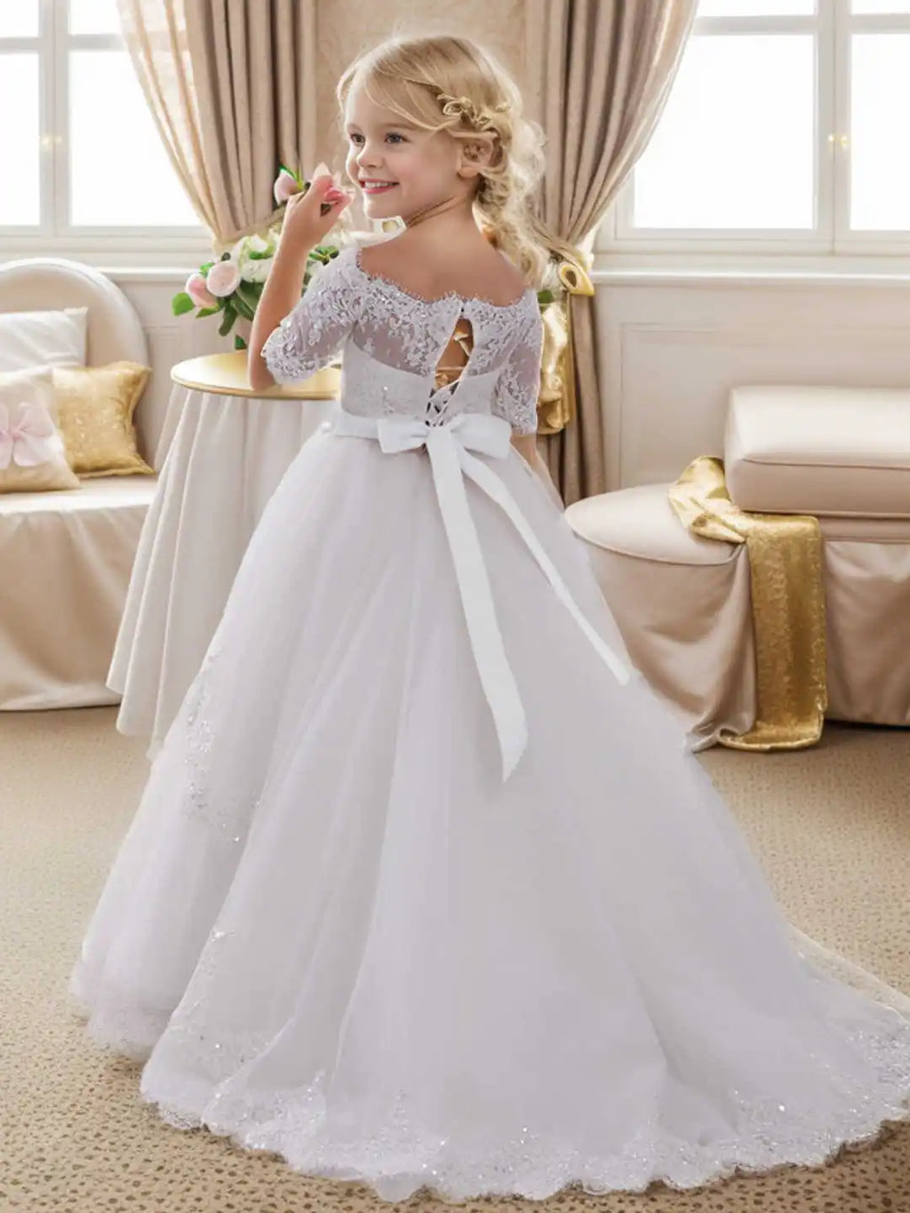 A girl in a white off-shoulder lace and tulle flower girl dress with a corset back and bow, standing in a room.