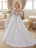 A girl in a white off-shoulder lace and tulle flower girl dress with a corset back and bow, standing in a room.