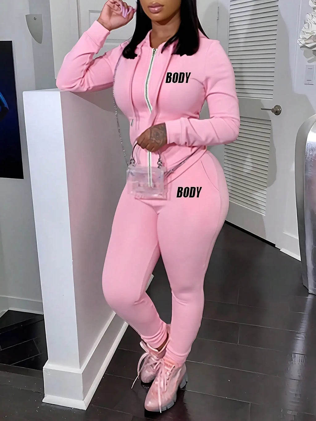 A woman wearing a pink two-piece set consisting of a zippered hoodie and sweatpants with black lettering.