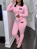 A woman wearing a pink two-piece set consisting of a zippered hoodie and sweatpants with black lettering.