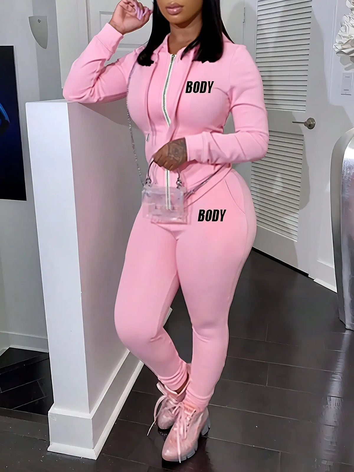 A woman wearing a pink two-piece set consisting of a zippered hoodie and sweatpants with black lettering.