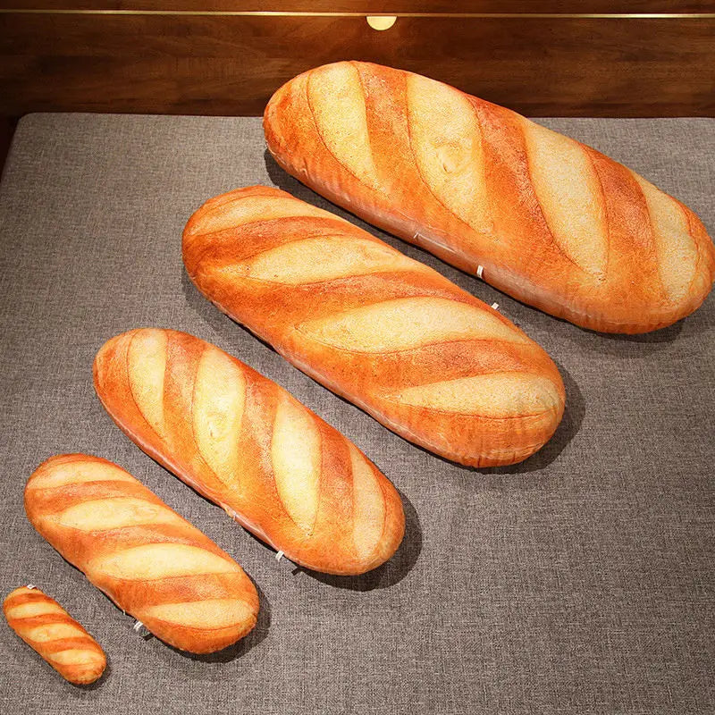 Five different sizes of realistic baguette-shaped plush pillows lined up in increasing order on a grey surface.