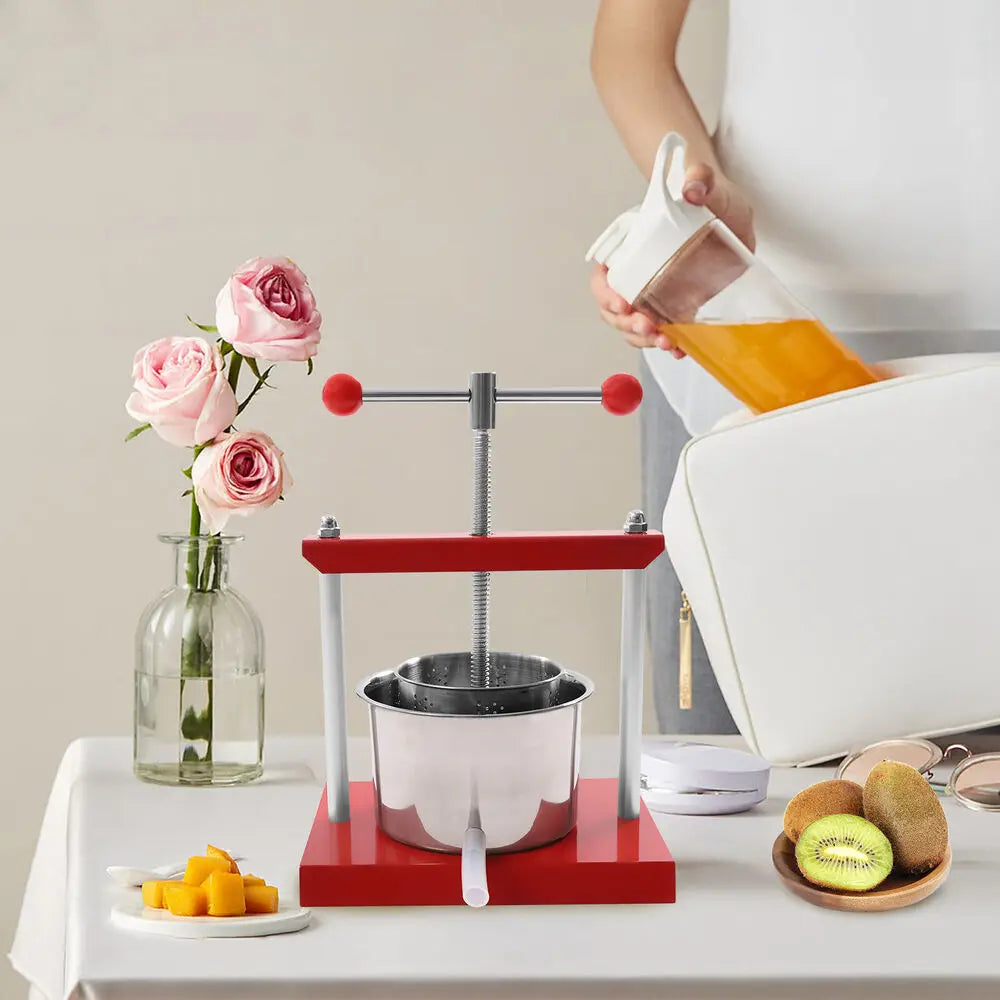 Manual fruit and herb press with stainless steel basket and red metal frame on a table with fruit and flowers