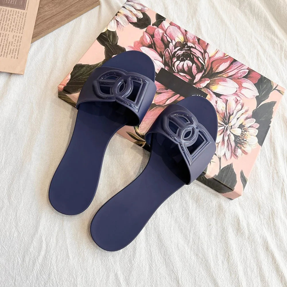 Pair of dark blue jelly slides with a decorative cutout letter emblem on the strap, resting on a floral box.