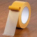 A roll of yellow grid-reinforced double-sided adhesive tape on a wooden surface.