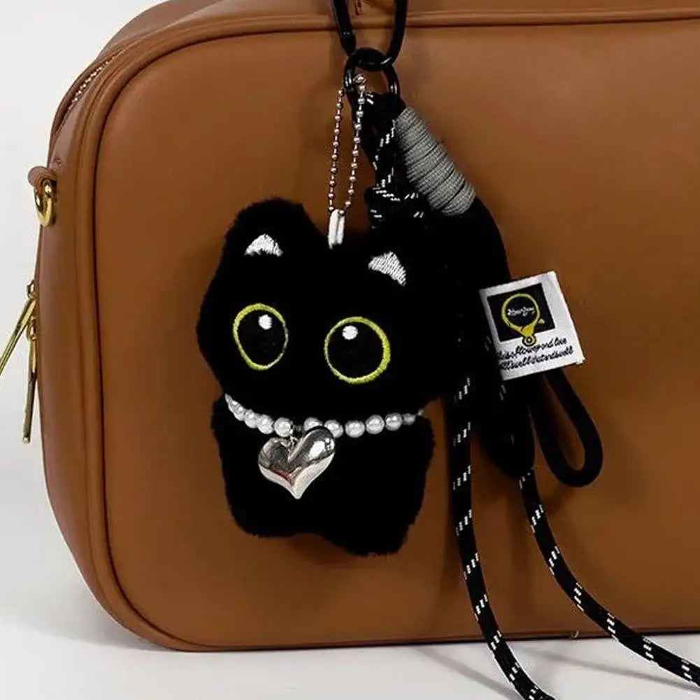 Black cat plush keychain with a pearl and heart necklace hanging on a brown bag.