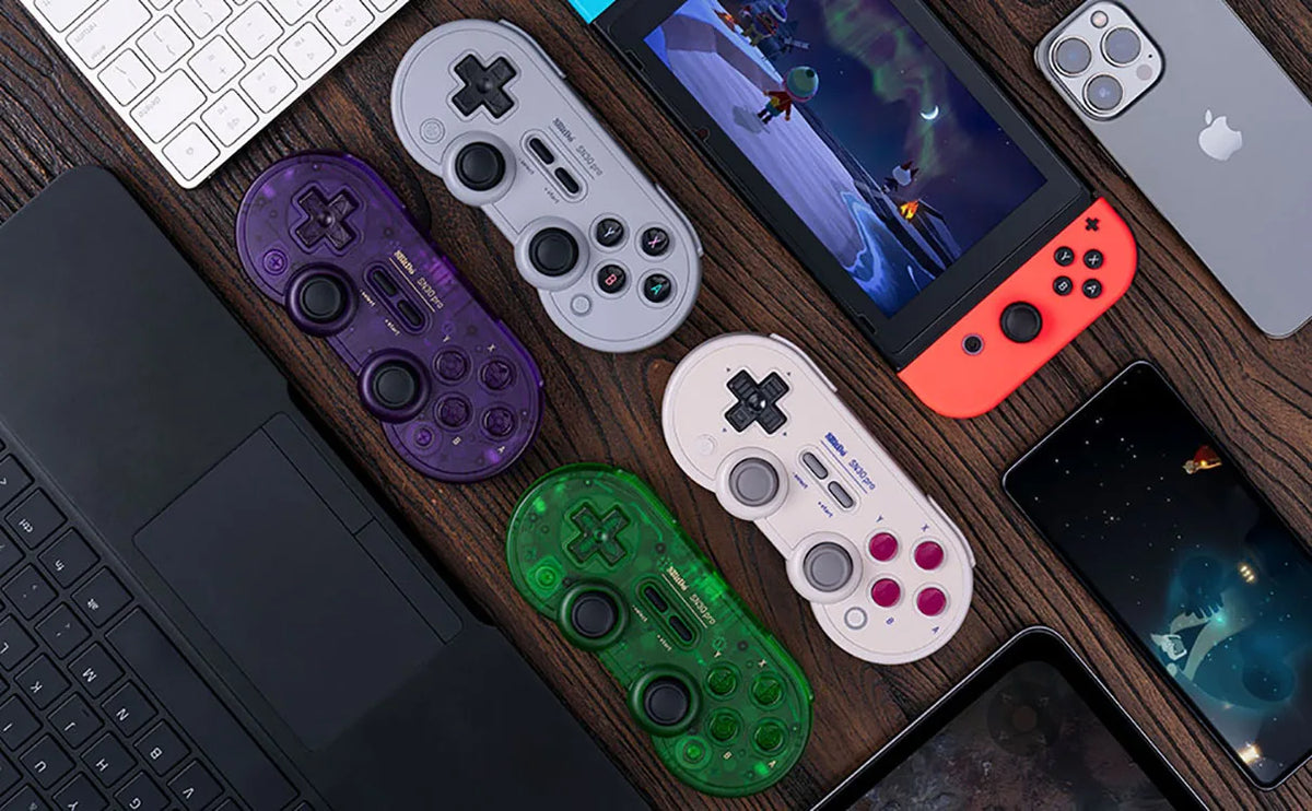 Top-down view of several game controllers, a keyboard, a portable gaming console, and smartphones on a wooden table.