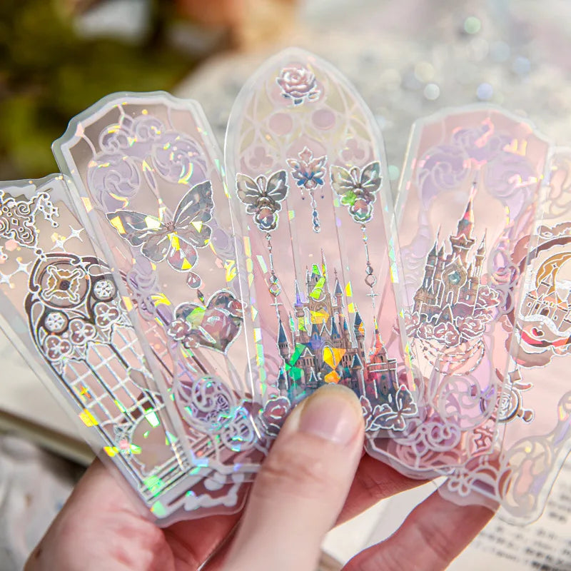 Hand holding several transparent holographic bookmarks featuring butterfly, castle, and rose designs.