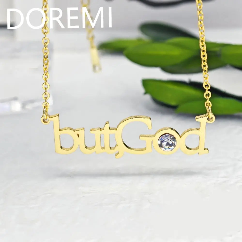 Gold necklace with the words butGod and a crystal on a light surface with green leaves