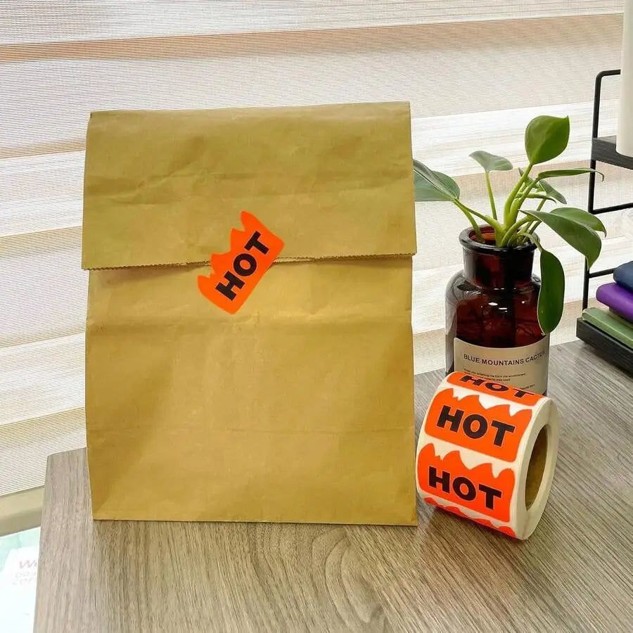 A brown paper bag with a flame-shaped HOT sticker and a roll of stickers next to a plant