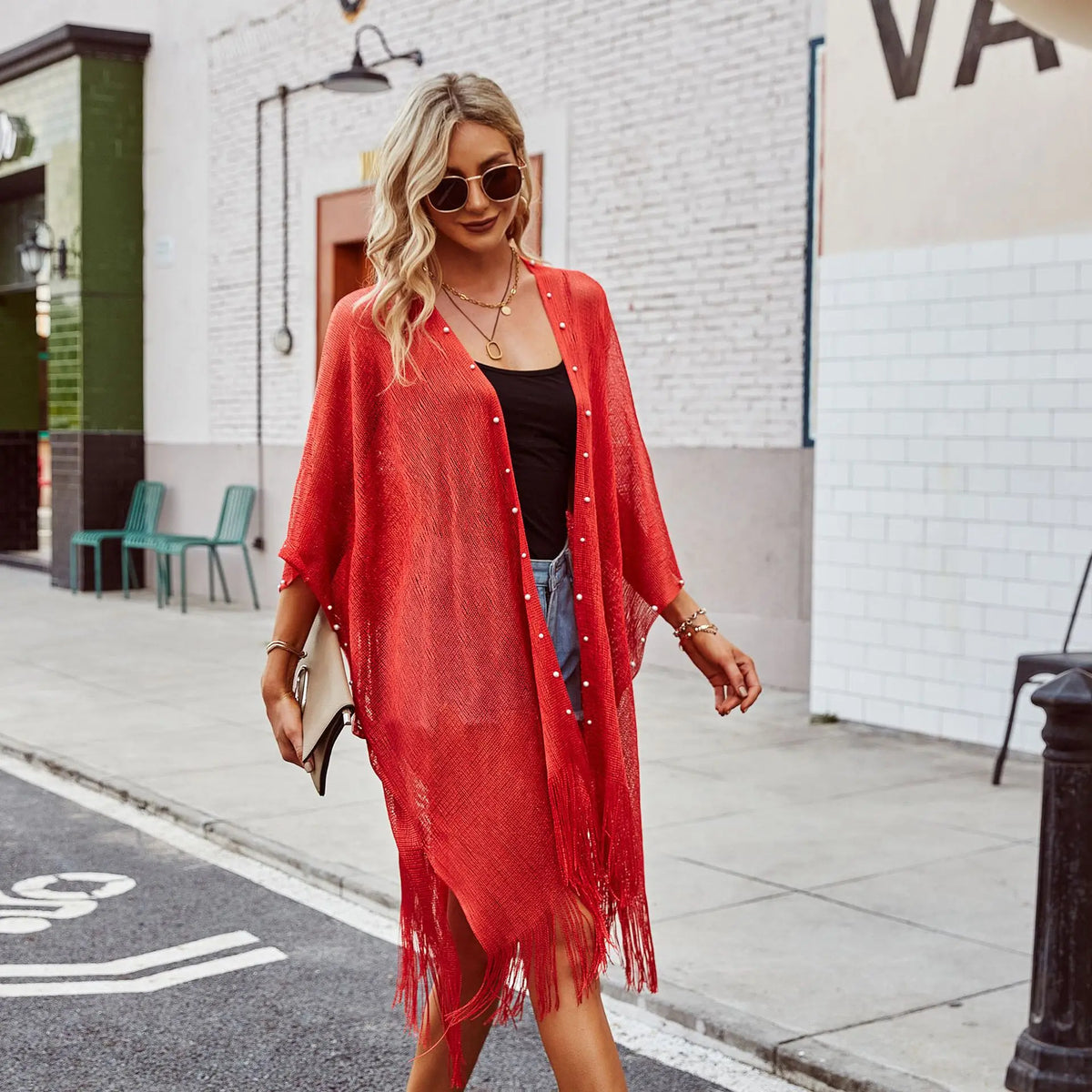 A woman wearing a red pearl-embellished fringe kimono cardigan over a black top and denim shorts.