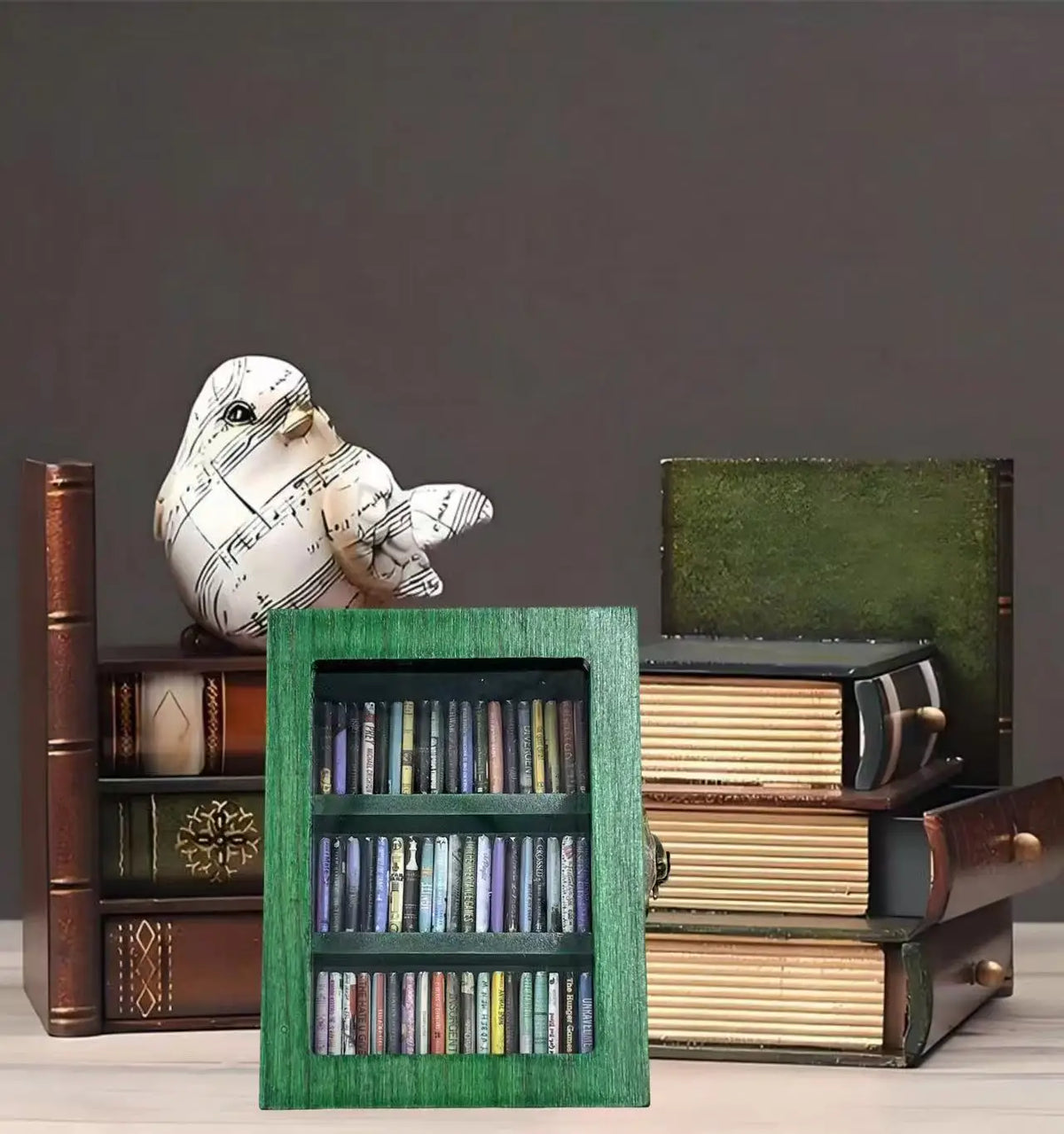 A small green wooden bookshelf filled with miniature books, displayed on a desk next to a decorative music-themed bird