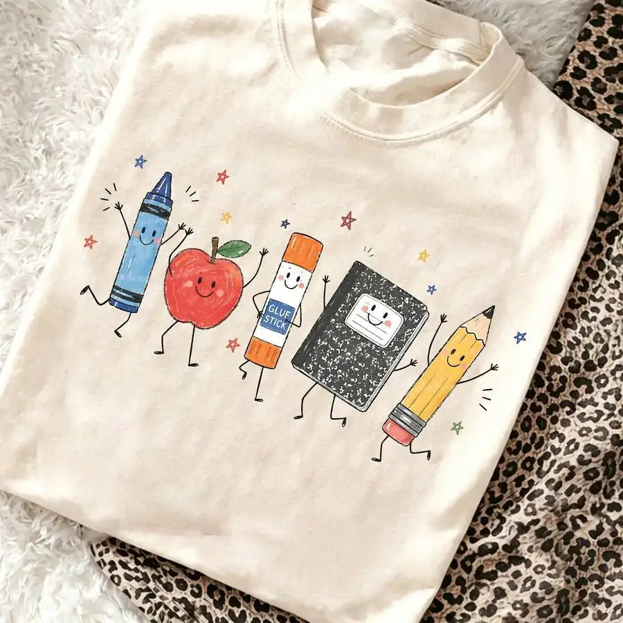 A cream-colored t-shirt featuring a cartoon illustration of smiling school supplies including a crayon, apple, glue stick