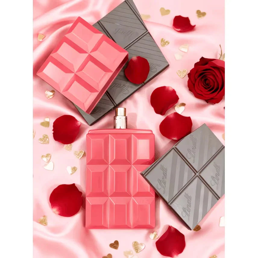 A perfume bottle shaped like a pink chocolate bar surrounded by chocolate squares, rose petals, and gold hearts.