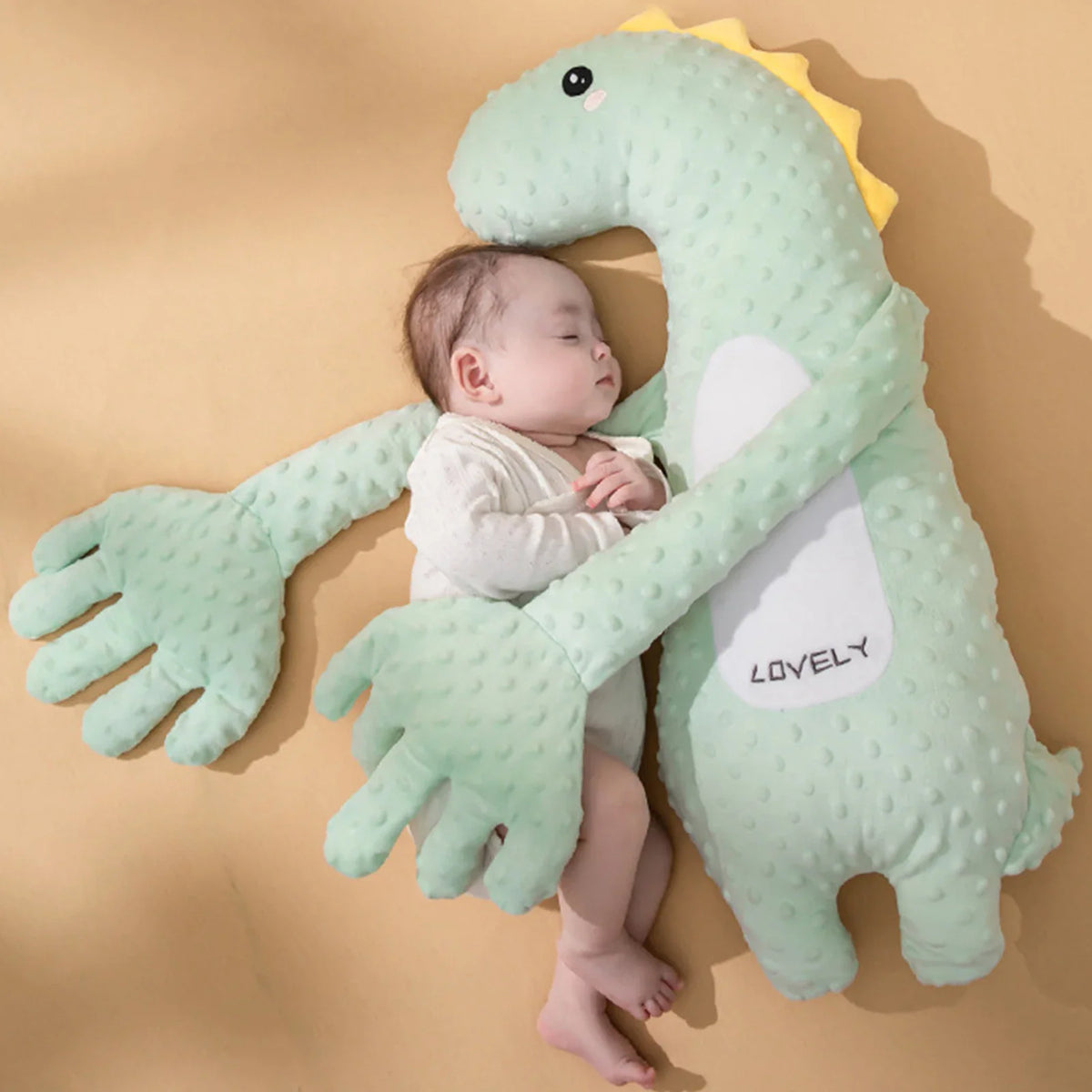 A light green plush dinosaur pillow with a baby sleeping next to it on a tan background.