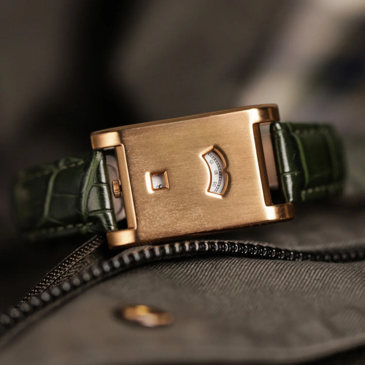 A rectangular rose gold watch with a green leather band resting on a dark fabric surface.