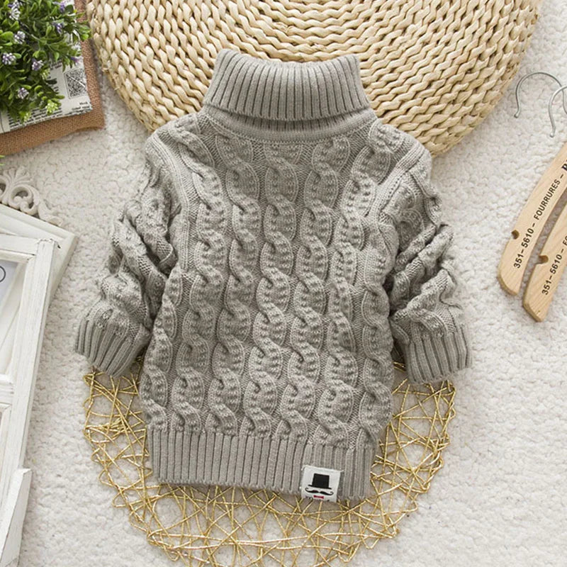 A grey cable-knit turtleneck sweater laid flat with a mustache logo patch.