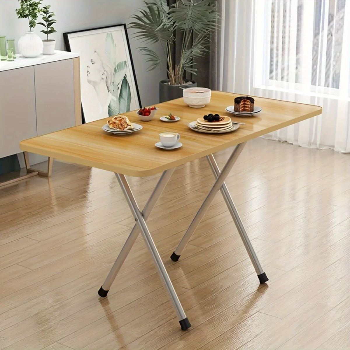 A small light-colored wood folding table with metal legs, set with breakfast food items, in a modern room.