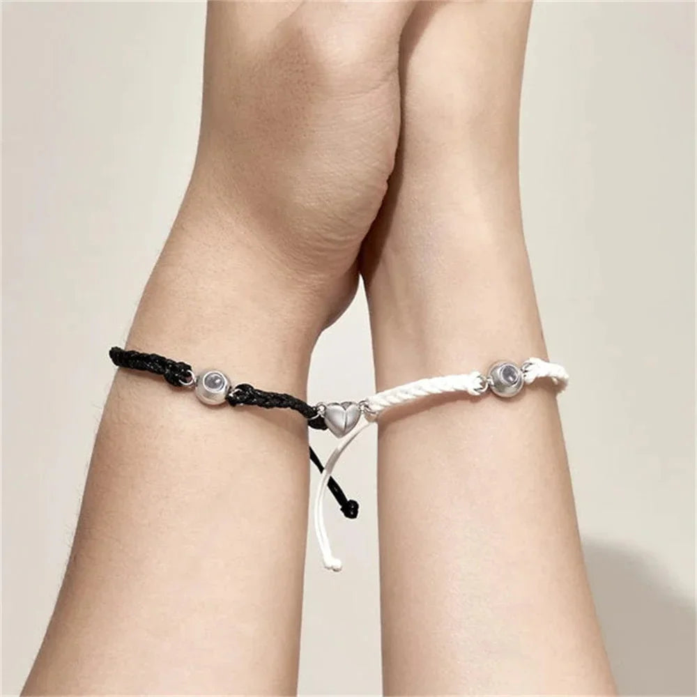 A pair of black and white braided adjustable bracelets with a magnetic heart clasp worn on wrists
