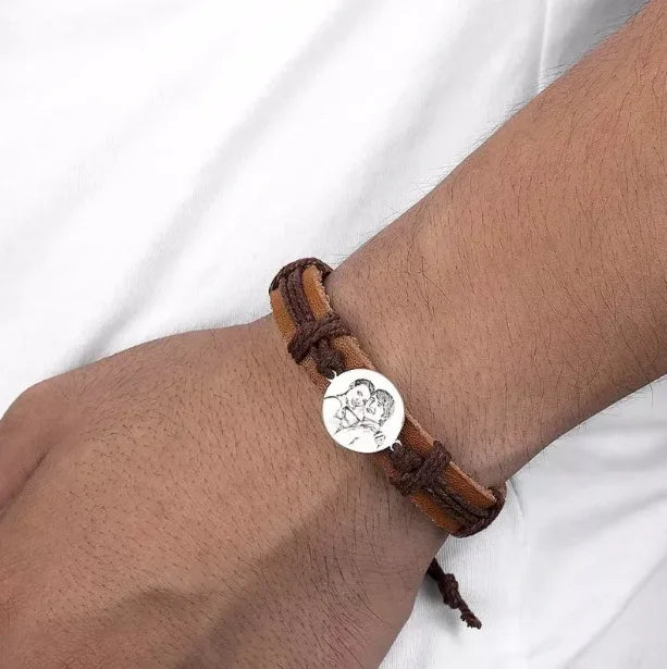 Men's leather bracelet with a round engraved metal charm worn on the wrist.
