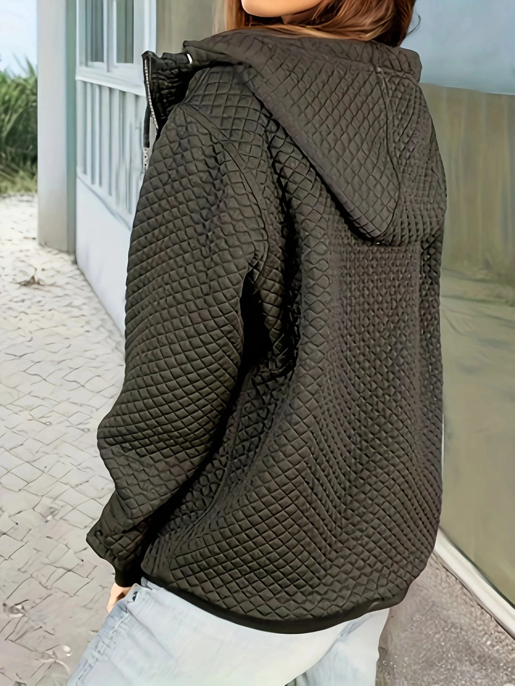 Rear view of a black diamond-quilted zip-up hooded jacket with ribbed cuffs, paired with blue jeans.