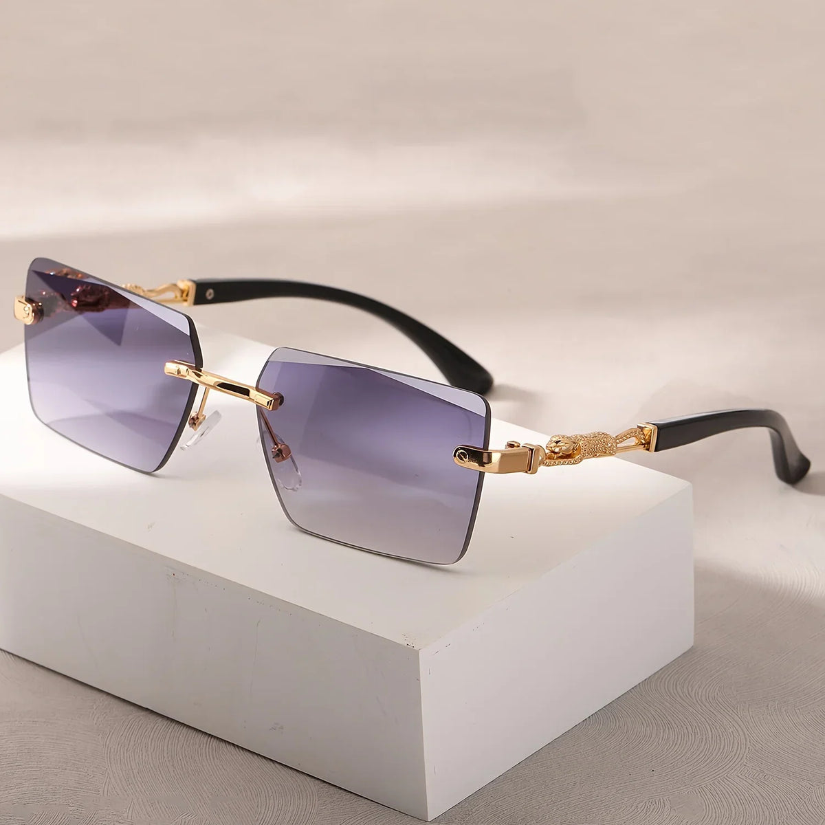 Rimless rectangular sunglasses with blue-grey gradient lenses and gold panther-style metal temples on a white block