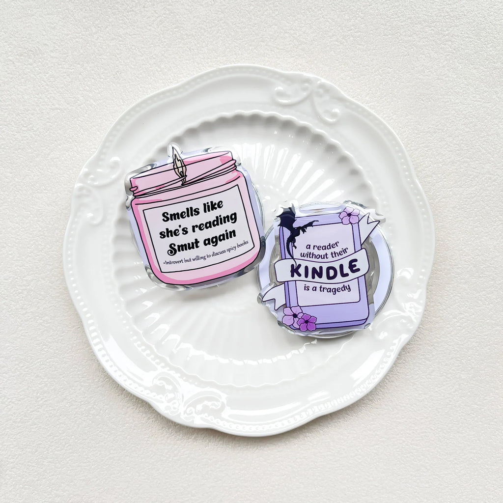 Two acrylic phone grips in candle and book designs resting on a white decorative plate.