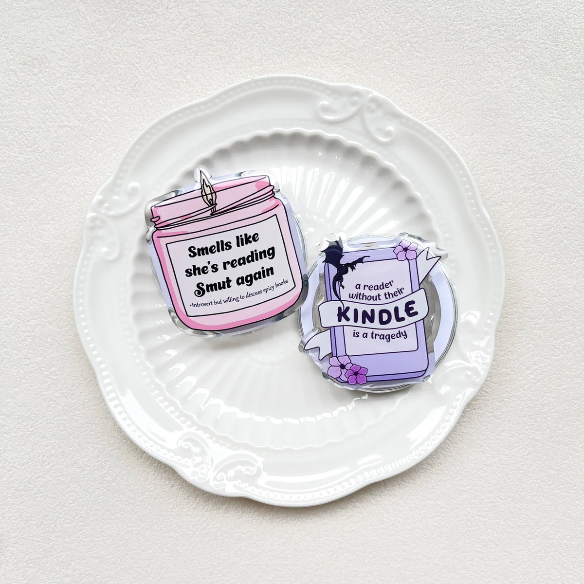 Two acrylic phone grips in candle and book designs resting on a white decorative plate.