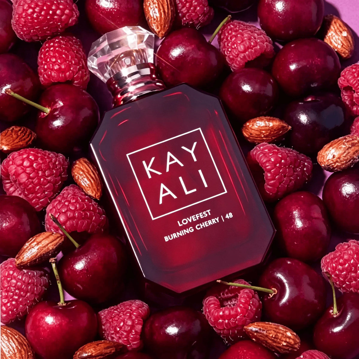 A red glass perfume bottle surrounded by scattered raspberries, cherries, and almonds.