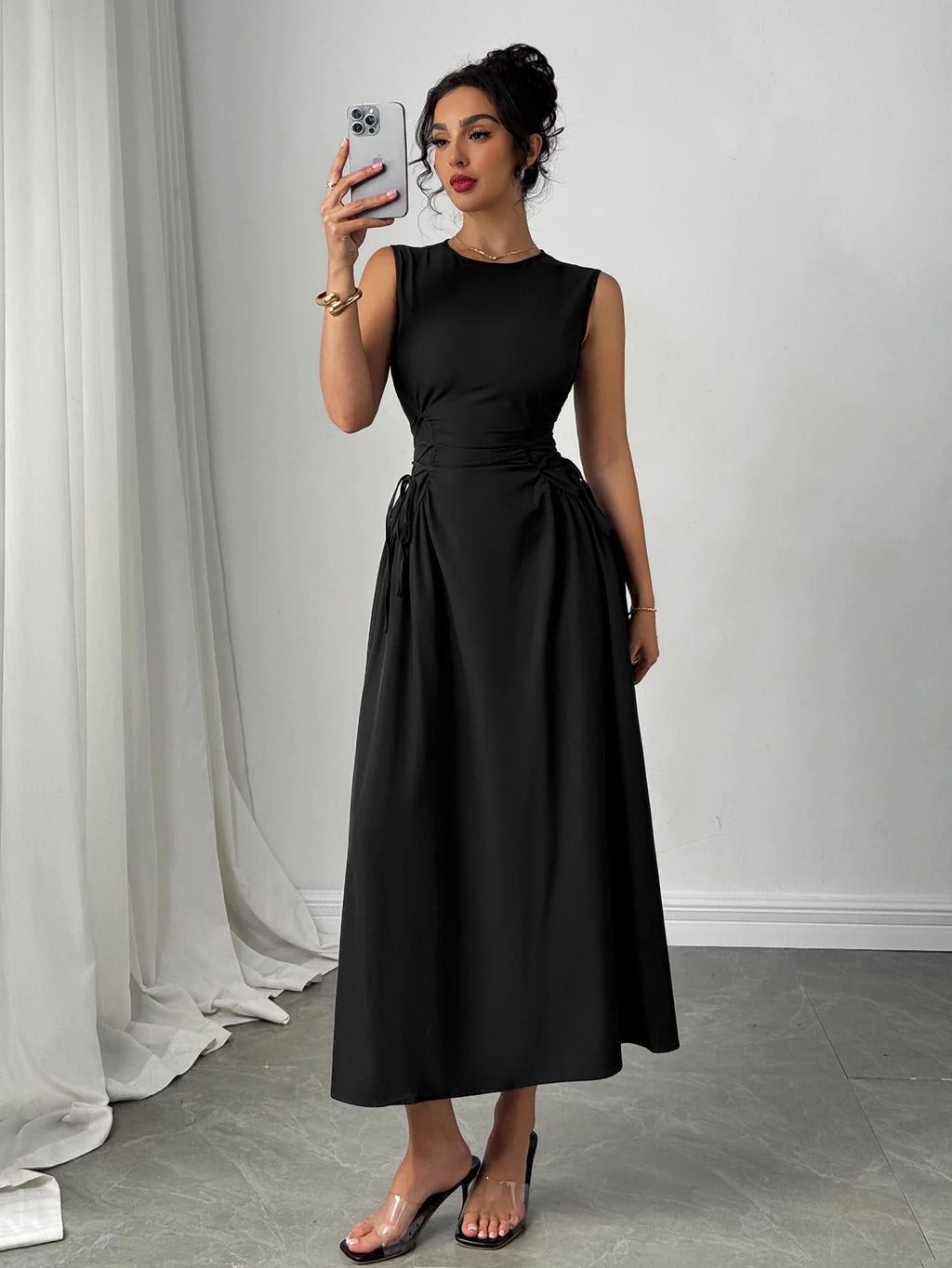 A woman posing in a black sleeveless midi dress while holding a phone to take a mirror selfie.