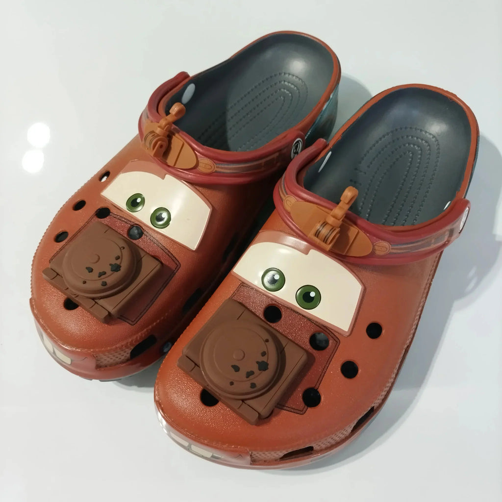 Front view of brown cartoon tow truck themed clogs with eyes and engine block charms.