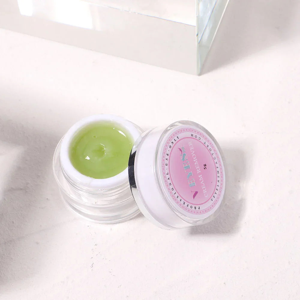 A small jar of green eyelash extension cream remover with the lid beside it on a white background.